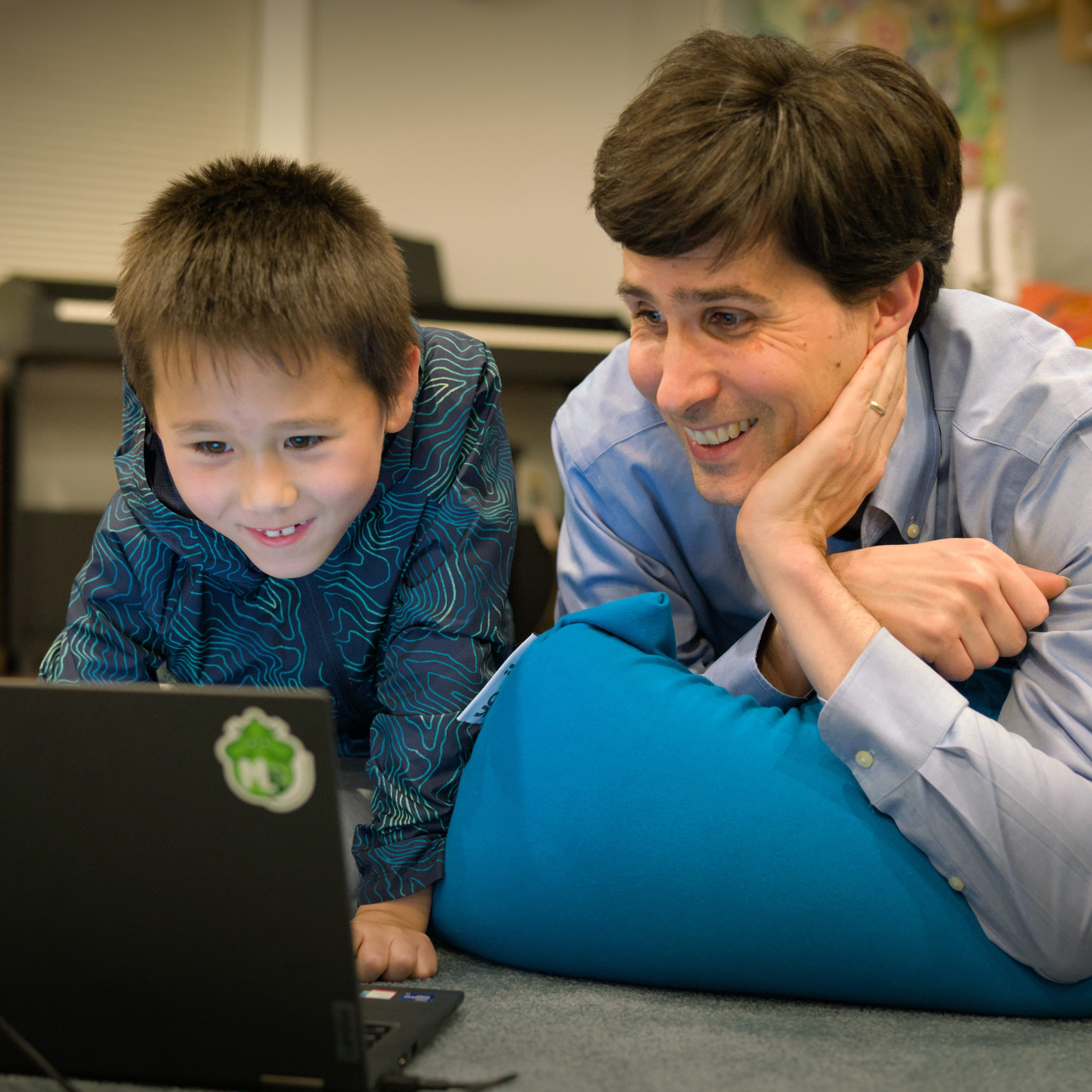 Son and father, laying down in front of a computer. A Music Blocks sticker is visible on the computer, an electronic keyboard is visible in the background.
